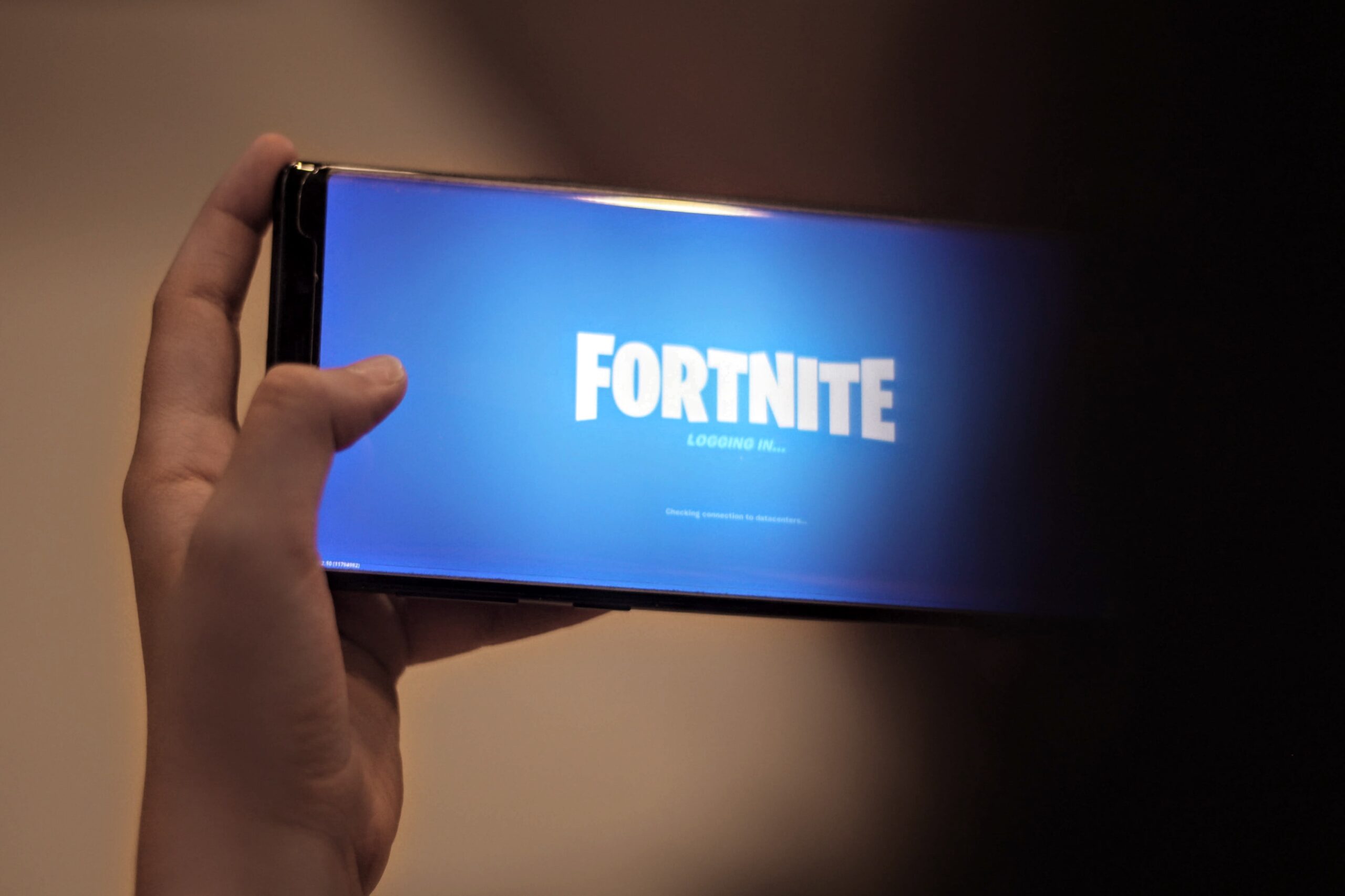 A hand holding a mobile phone sideways, showing the Fortnite logo on a loading screen.