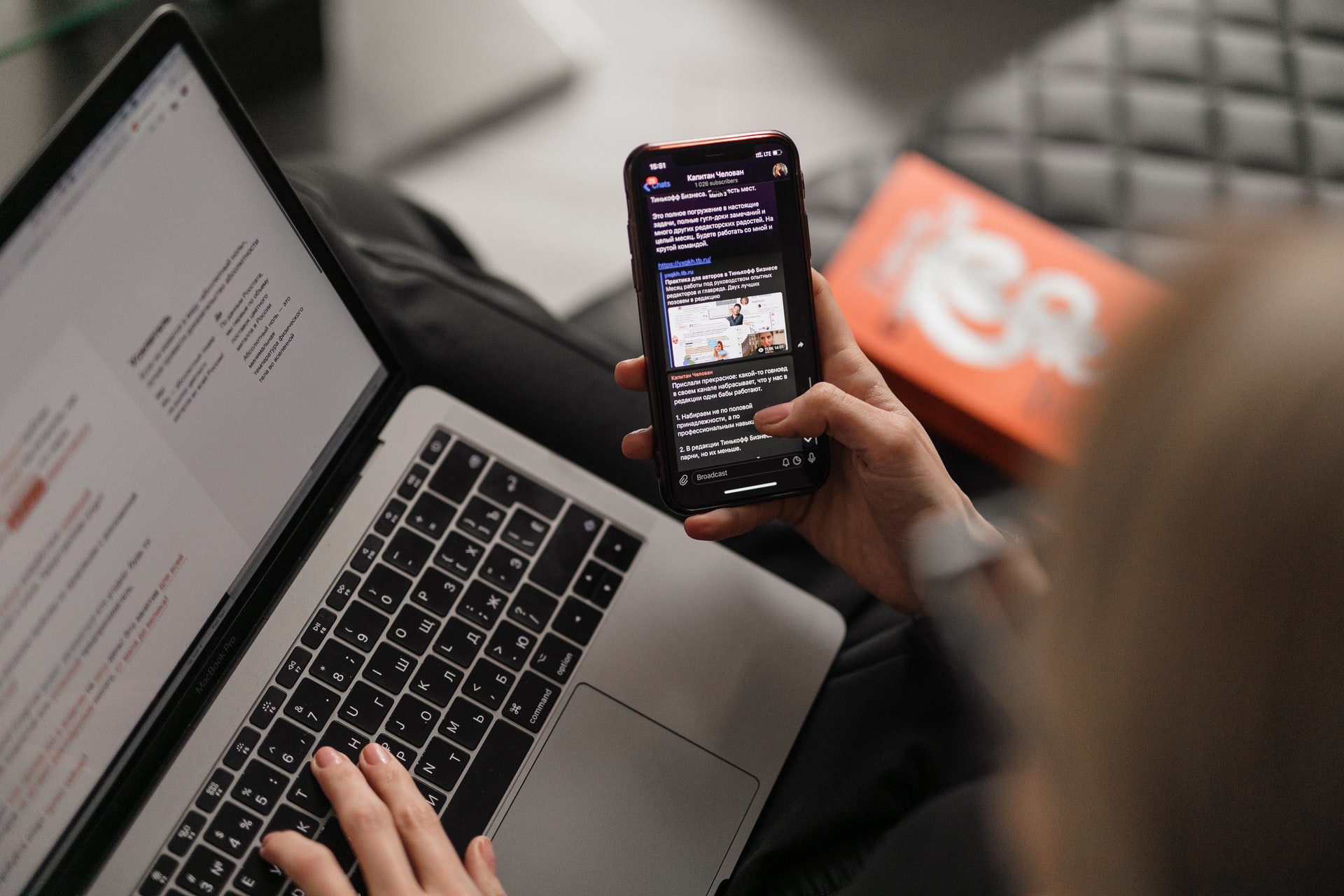 A person looking at a mobile phone in one hand while typing on a laptop.