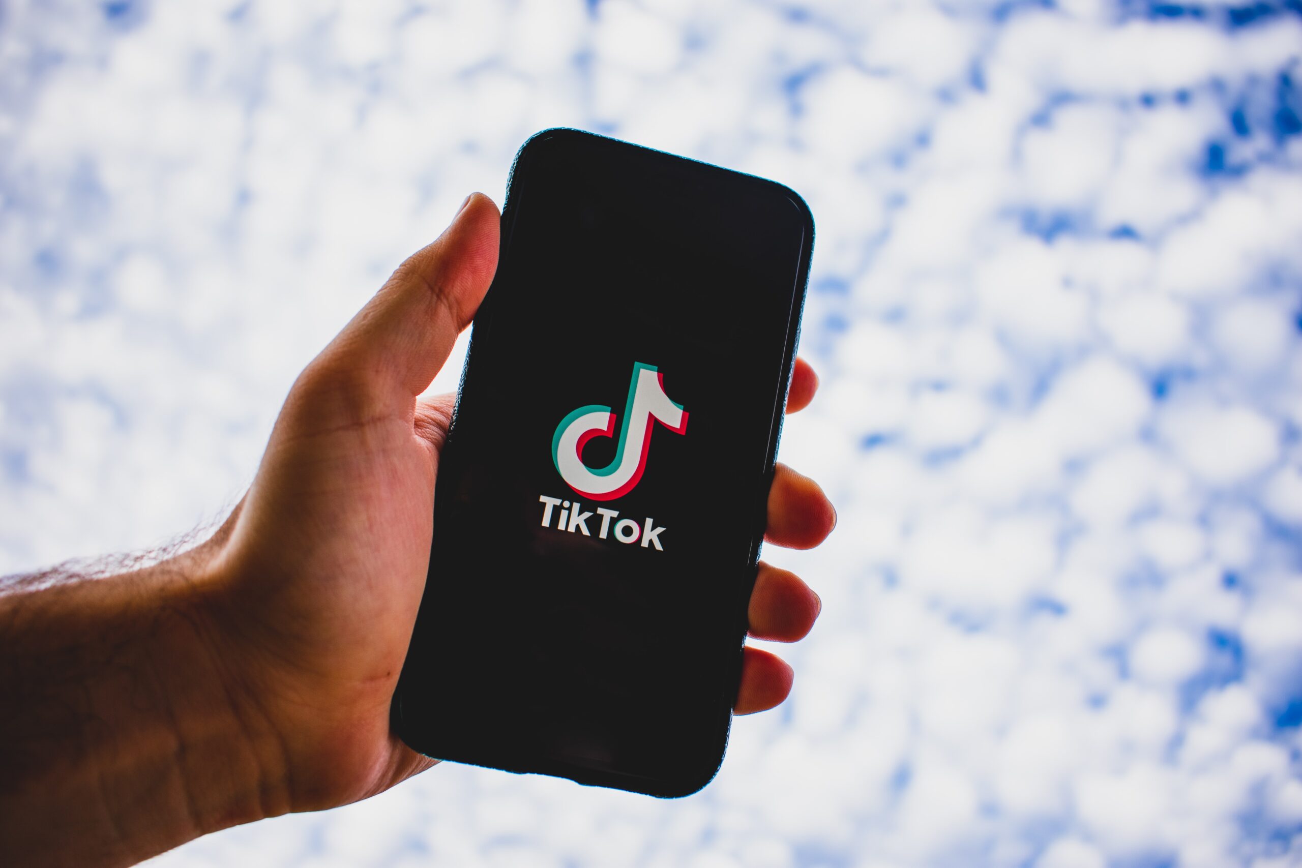 A hand holding up a mobile phone showing the TikTok logo against a cloudy blue sky.